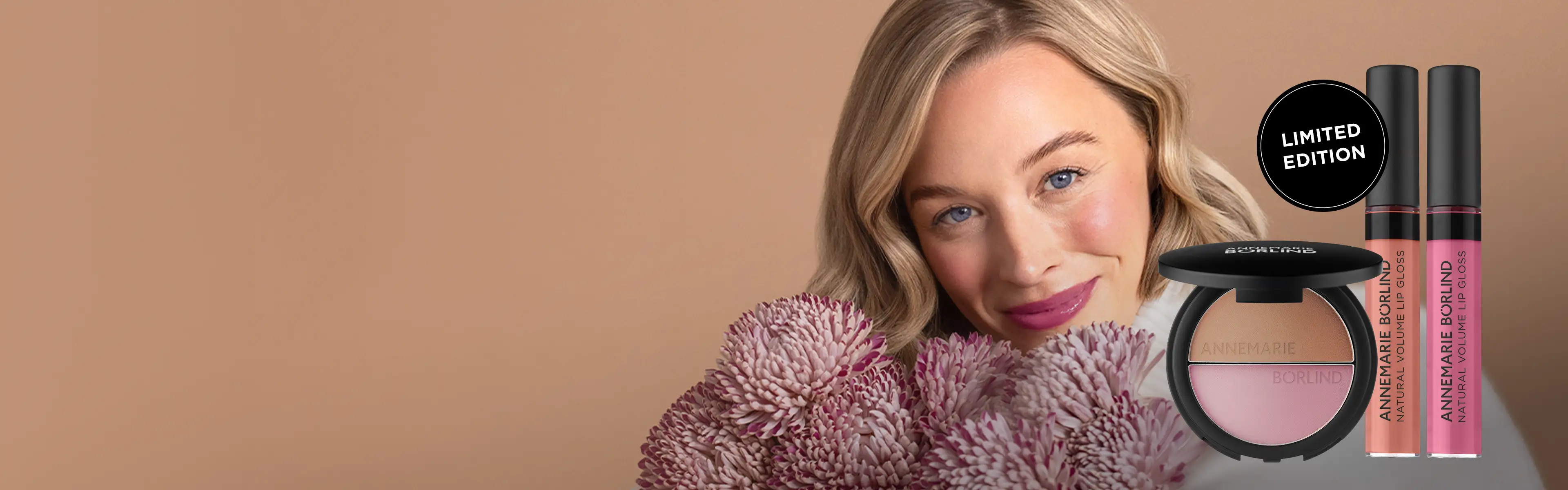 Frau mit ANNEMARIE BÖRLIND NATURAL HUG SET Make-up, lehnt sich lächelnd mit Blumenstrauß in Rosa- und Lilatönen auf Tisch vor beigem Hintergrund