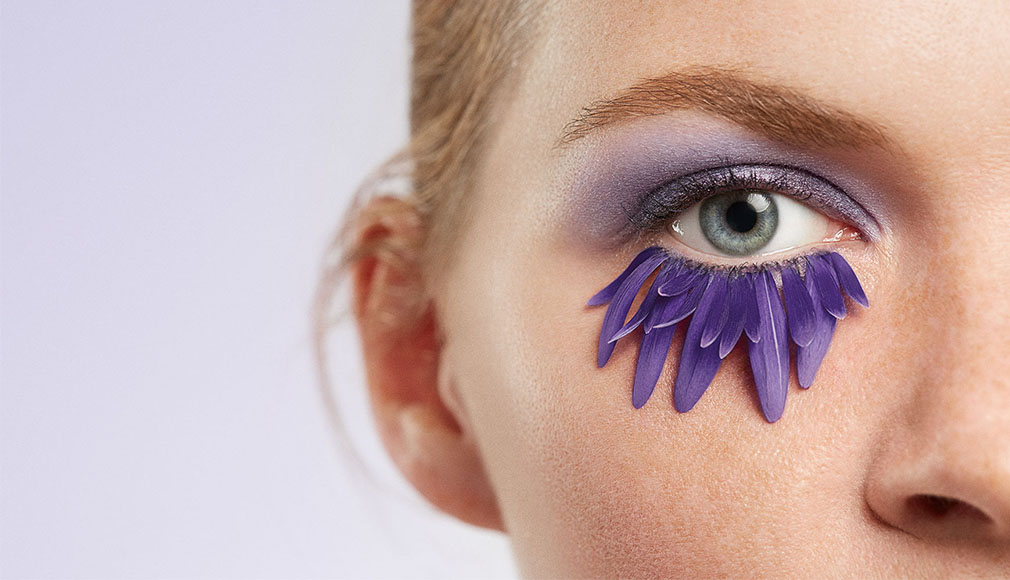 Nahaufnahme eines Auges mit violettem Lidschatten und künstlichen violetten Blütenblättern als untere Wimpernverlängerung