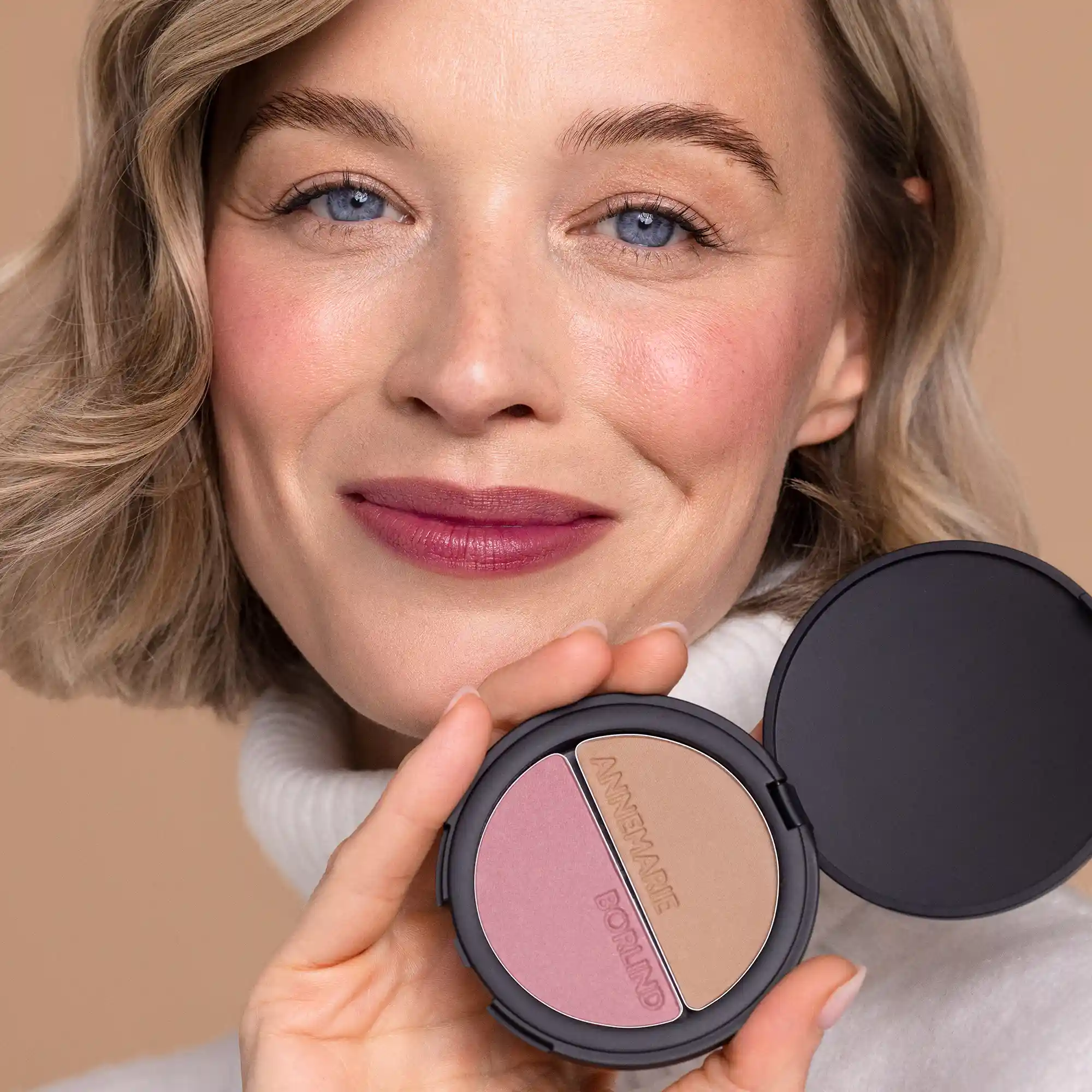 Frau mit frischem Teint hält ANNEMARIE BÖRLIND 2IN1 BRONZER & SOFT BLUSH in schwarzer Dose vor beigem Hintergrund