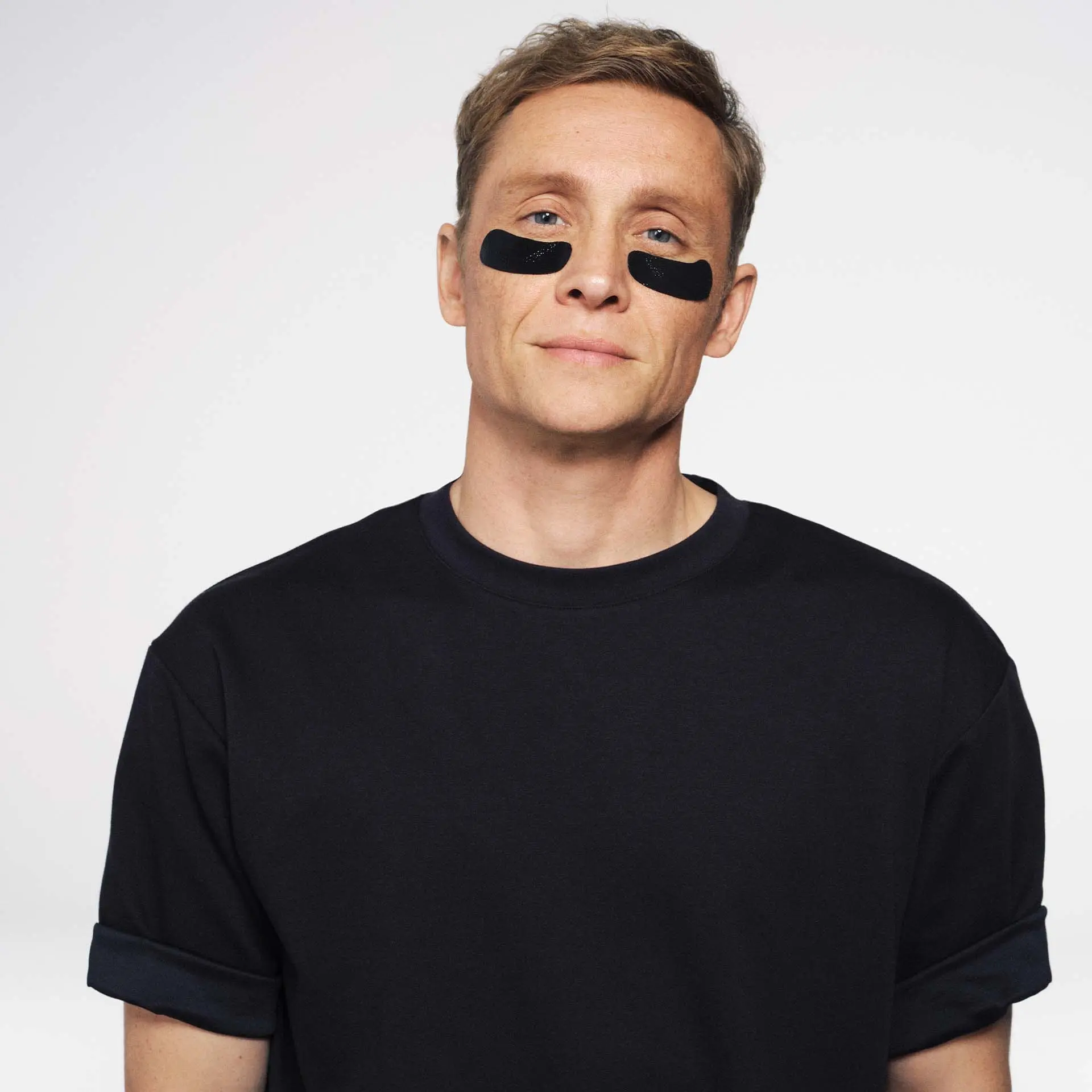Matthias Schweighöfer mit schwarzen Augenpads unter den Augen vor hellem Hintergrund, trägt schwarzes T-Shirt