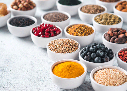 Verschiedene Superfoods wie Kurkuma, Blaubeeren, Granatapfelkerne, Nüsse und Samen in weißen Schalen auf hellem Untergrund