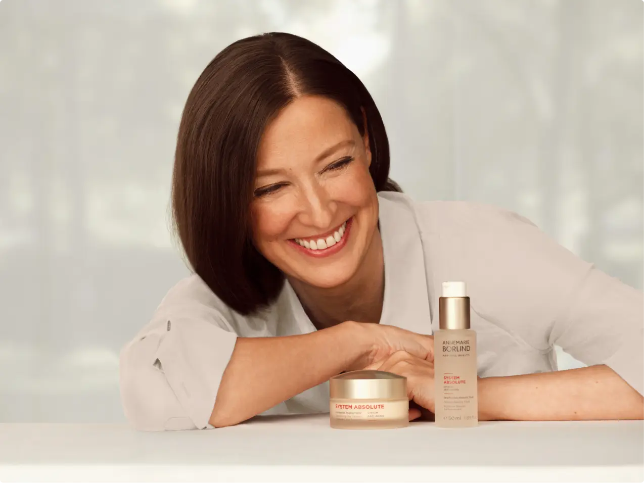 Frau lächelt und lehnt sich auf Tisch mit ANNEMARIE BÖRLIND SYSTEM ABSOLUTE Creme und Serum