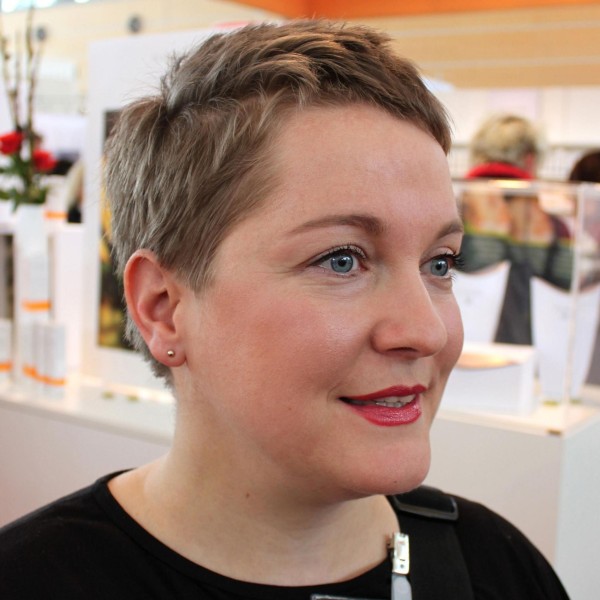 Nahaufnahme einer Frau mit kurzem Haar und dezentem Make-up, im Hintergrund Kosmetikprodukte sichtbar