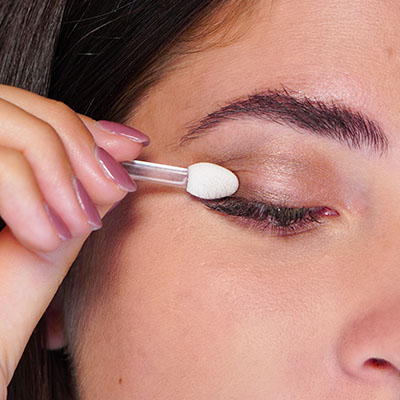 Nahaufnahme einer Frau, die mit einem Applikator hellen Lidschatten auf das geschlossene Augenlid aufträgt.