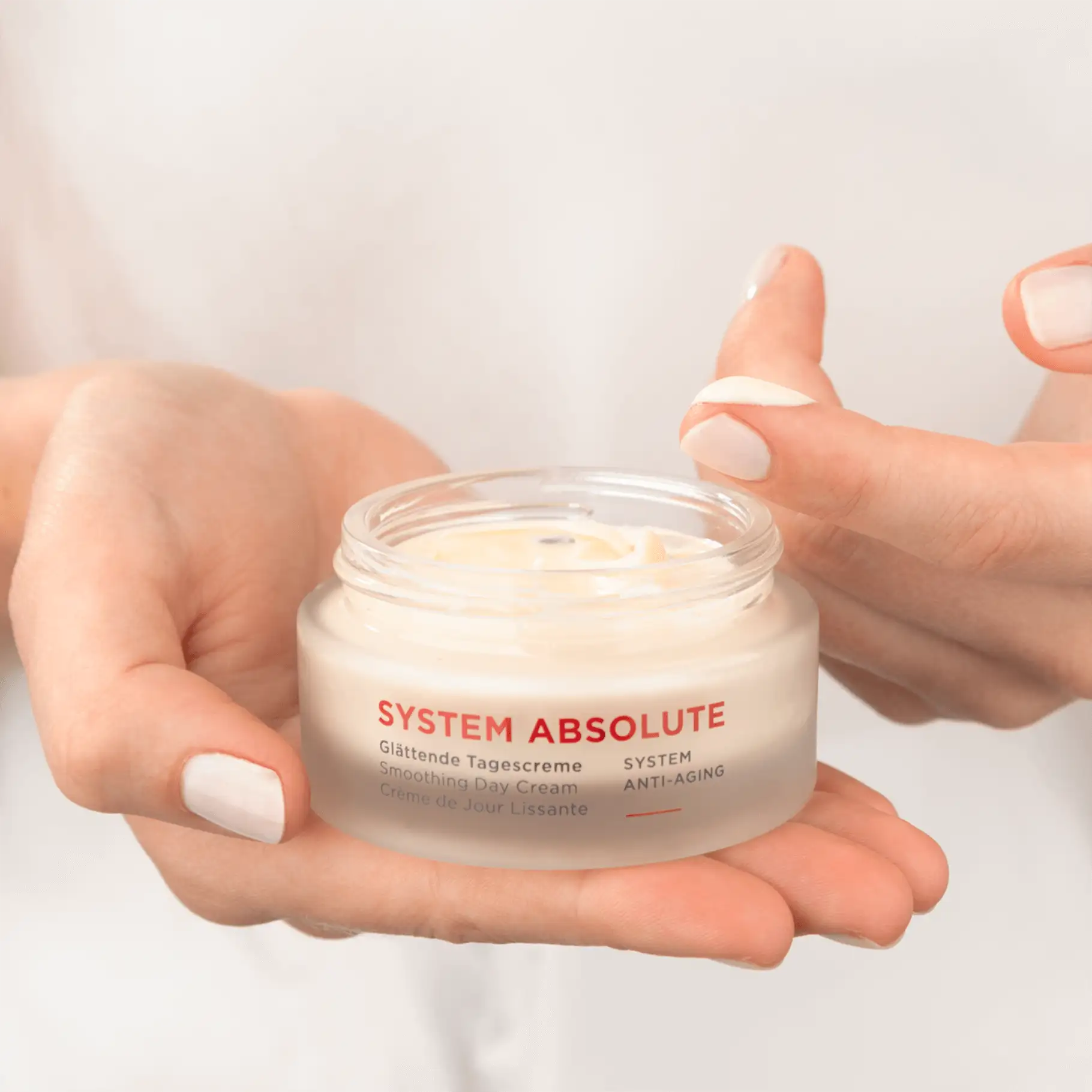 SYSTEM ABSOLUTE Glättende Tagescreme im Tiegel, Hand hält Creme, Finger nimmt Produktprobe