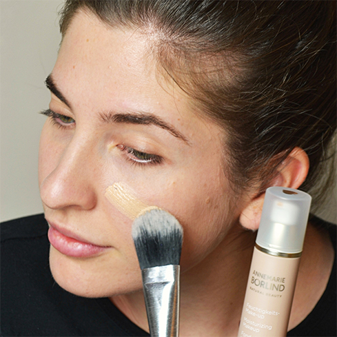 Frau trägt mit Pinsel flüssige Foundation von ANNEMARIE BÖRLIND auf die Wange auf