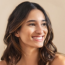 Nahaufnahme einer lächelnden Frau mit glatter, strahlender Haut und braunen, lockigen Haaren vor hellem Hintergrund