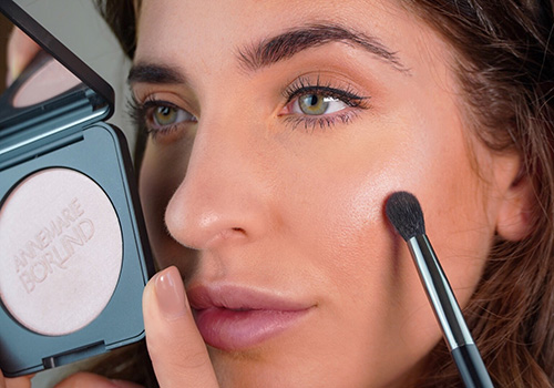 Nahaufnahme einer Frau, die ANNEMARIE BÖRLIND Coral Glow Highlight-Puder mit Pinsel auf die Wange aufträgt