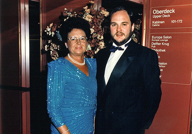 Annemarie Lindner und Sohn Michael in eleganter Abendkleidung stehen vor einem Schild mit Deck- und Kabineninformationen auf einem Schiff