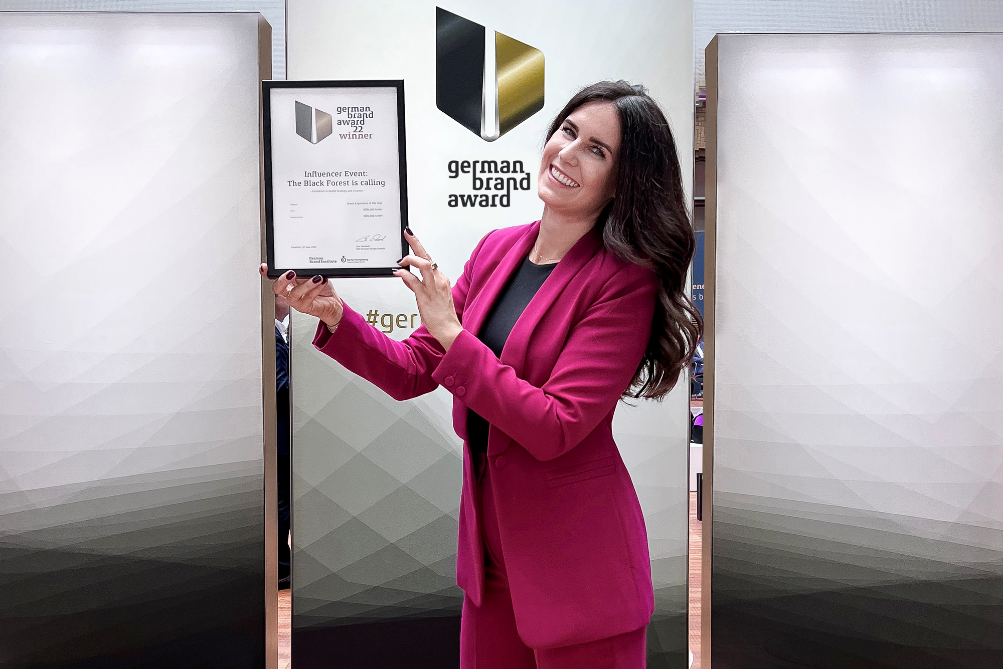 Frau in pinkem Anzug hält Urkunde des German Brand Award 2022 vor Logo und lächelt in die Kamera