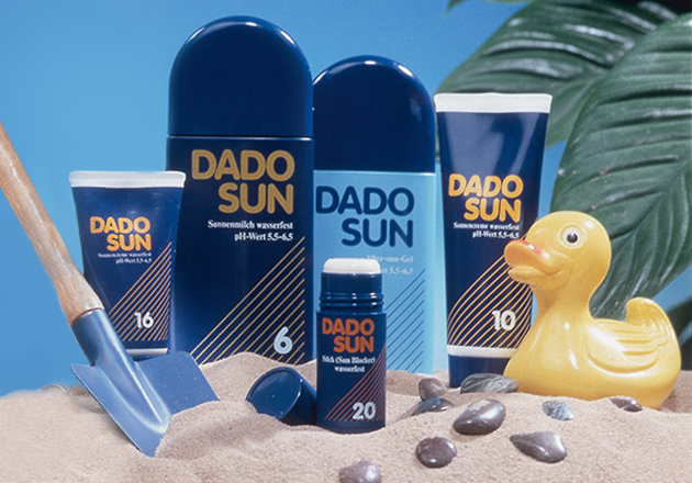 DADO SUN Sonnenmilch und -gel in verschiedenen LSF, Sand, gelbe Badeente und blaue Schaufel im Sand