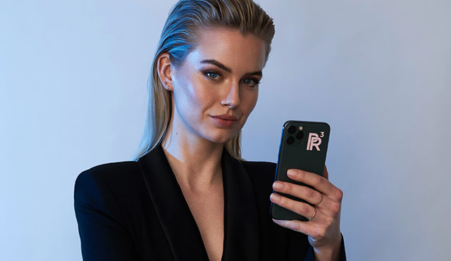 Frau mit schwarzem Blazer hält Smartphone mit Aufkleber "PR³" vor blauem Hintergrund