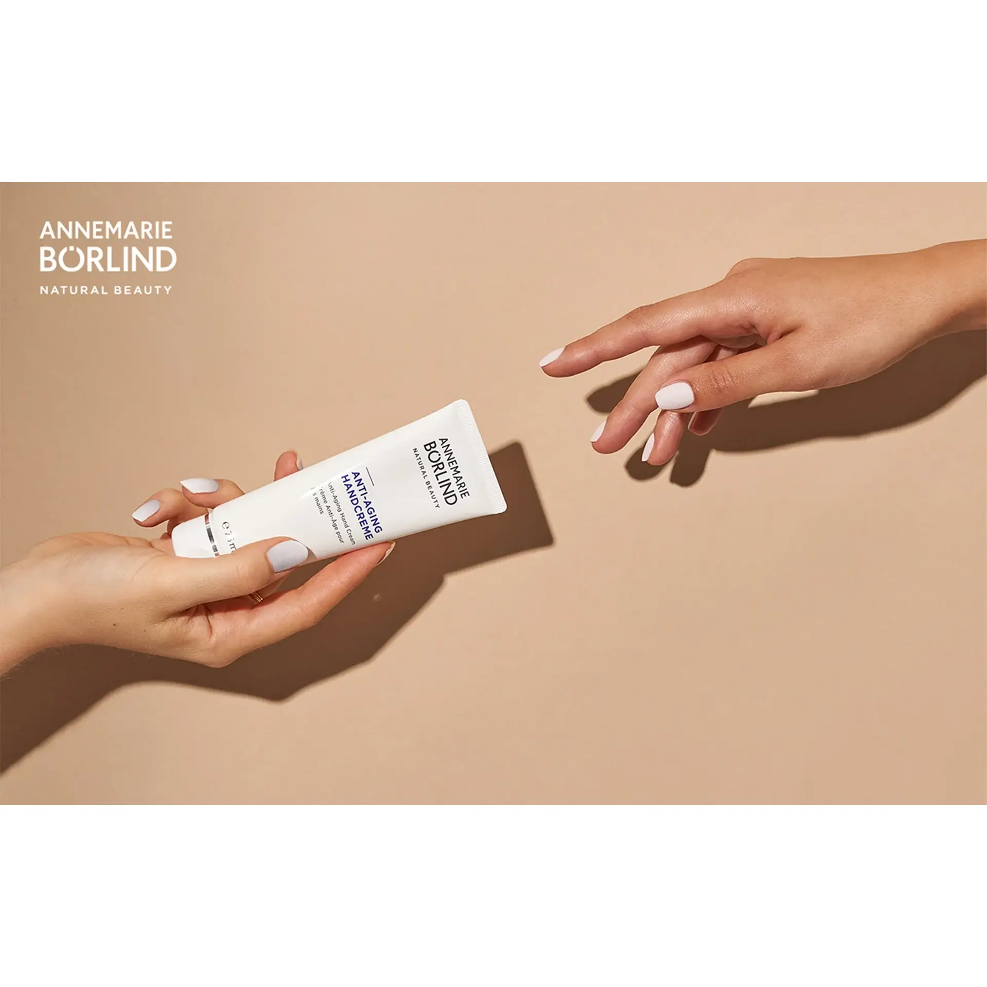Geschenkgutschein ANNEMARIE BÖRLIND ANNEMARIE BÖRLIND Anti-Aging Handcreme, eine Hand reicht die Tube an eine andere Hand mit beigem Hintergrund
