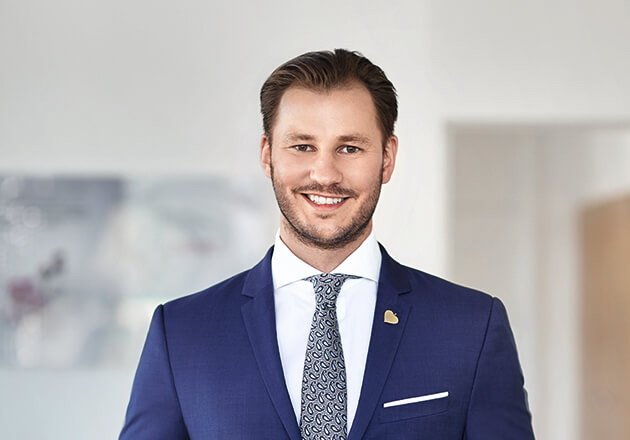 Mann in blauem Anzug und gemusterter Krawatte lächelt in einem hellen, unscharfen Raum