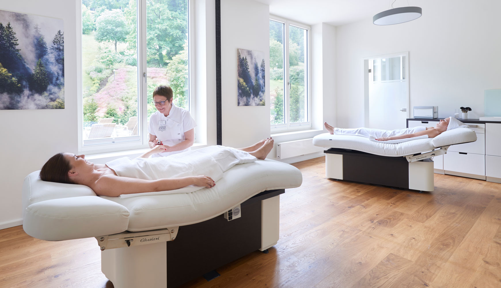 Zwei Frauen liegen auf Behandlungsliegen in hellem Spa-Raum, eine Kosmetikerin behandelt Hände einer Kundin.