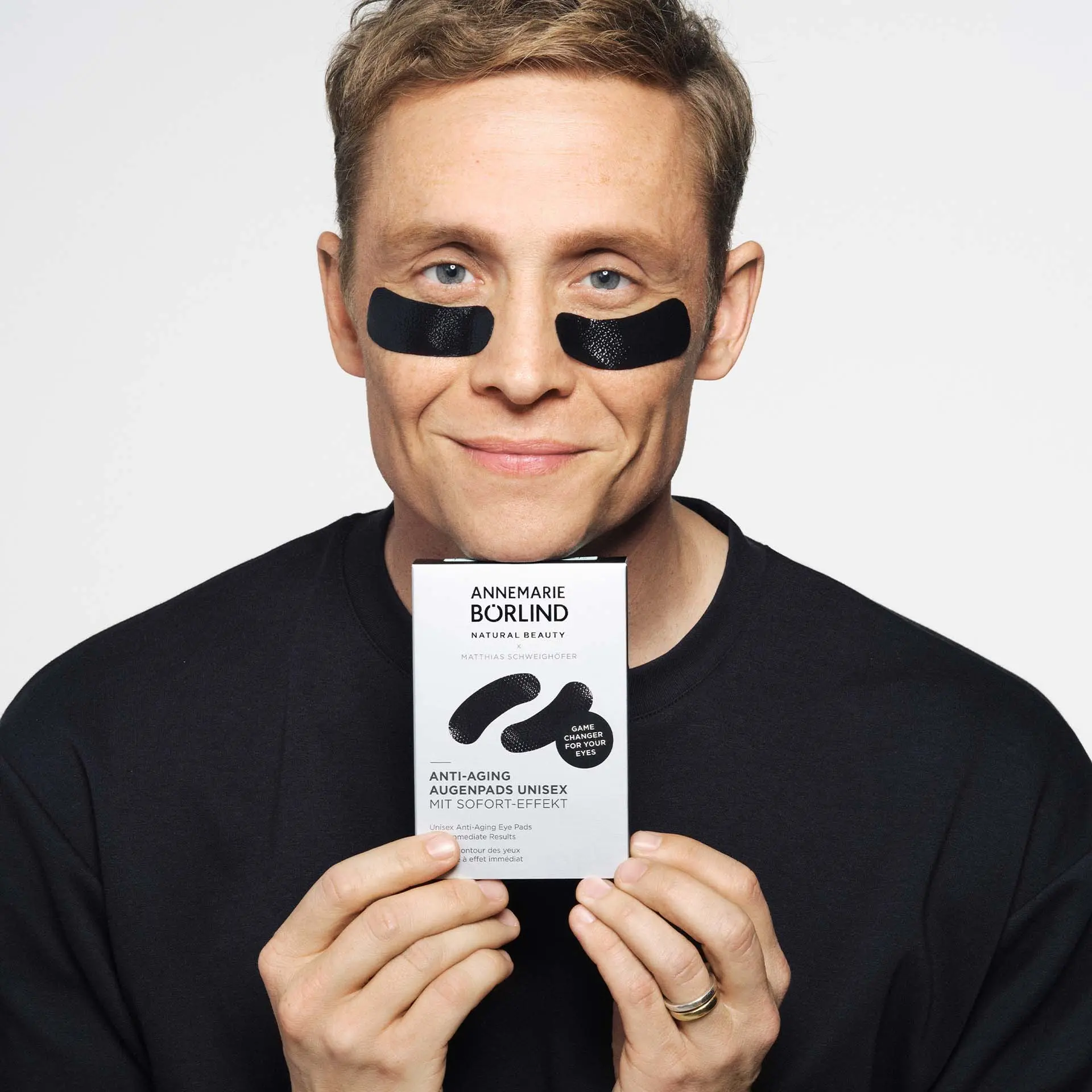 Mann mit schwarzen ANNEMARIE BÖRLIND Anti-Aging Augenpads unter den Augen hält Produktverpackung vor weißem Hintergrund