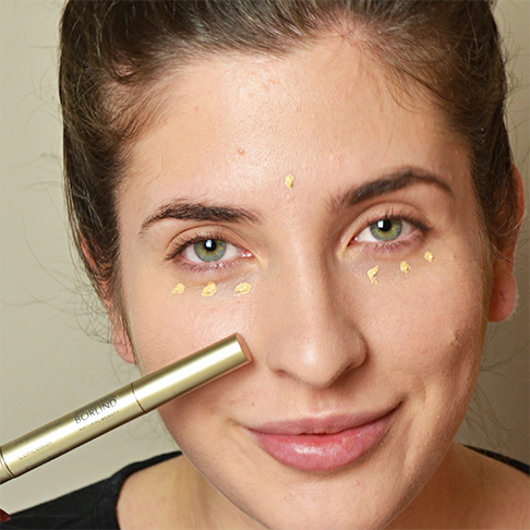 Frau trägt goldfarbenen Concealer punktuell unter den Augen und auf der Stirn auf, Nahaufnahme