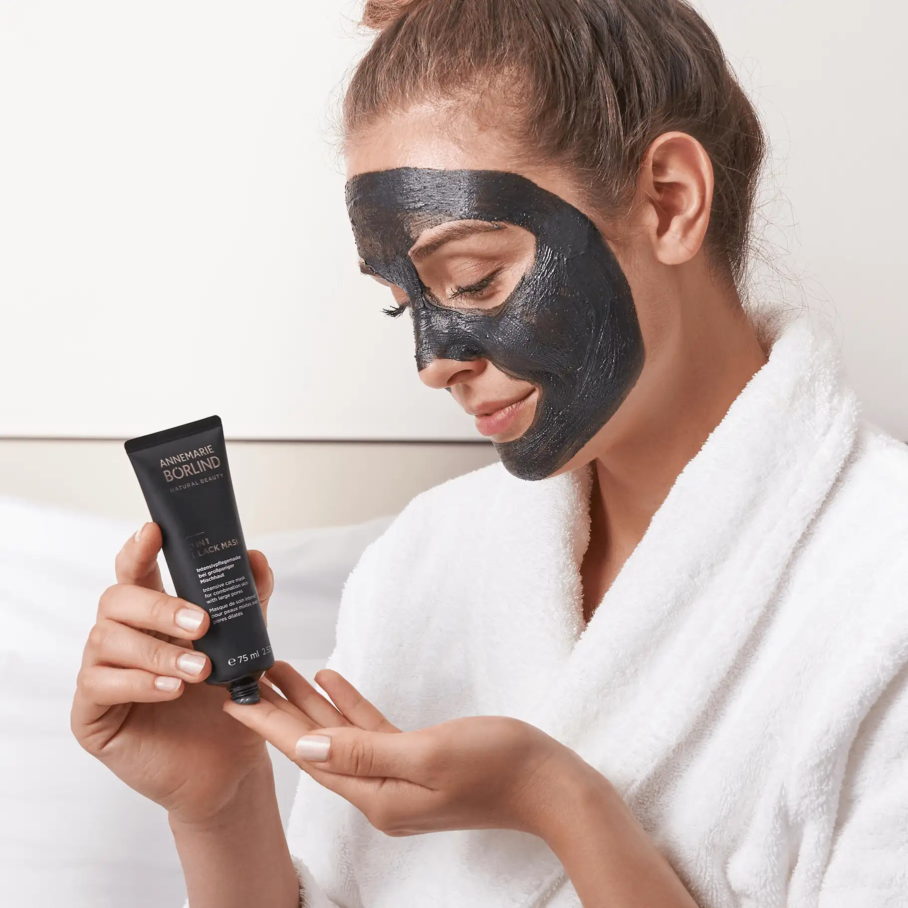 Frau mit schwarzer Gesichtsmaske trägt ANNEMARIE BÖRLIND 2-IN-1 BLACK MASK auf Finger auf, weißer Bademantel