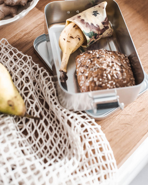 Brotdose mit Vollkornbrötchen und Banane in Bienenwachstuch, daneben ein Netz-Einkaufsbeutel auf Holzoberfläche