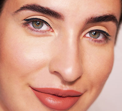 Nahaufnahme eines Gesichts mit grünlichen Augen, schwarzem Lidstrich und mattem, terrakottafarbenem Lippenstift