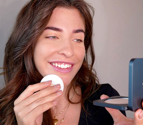 Frau trägt mit Schwämmchen Puder auf, lächelt in Kompaktspiegel, natürliche Make-up-Situation