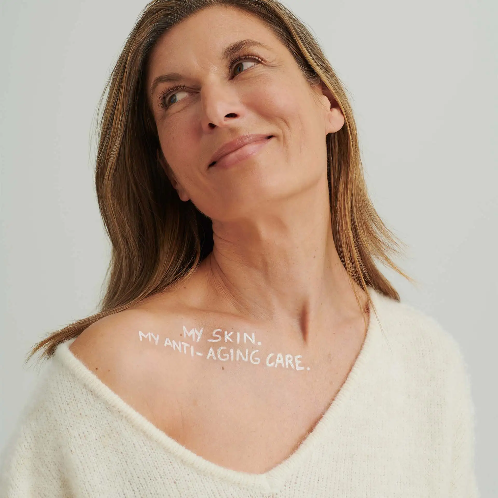 Frau mit offenem Pullover zeigt Schulter mit Schriftzug „MY SKIN. MY ANTI-AGING CARE.“ auf heller Haut