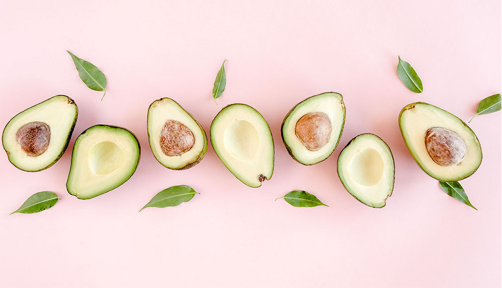 Mehrere halbierte Avocados mit grünen Blättern auf rosa Hintergrund, Fruchtfleisch und Kerne sichtbar