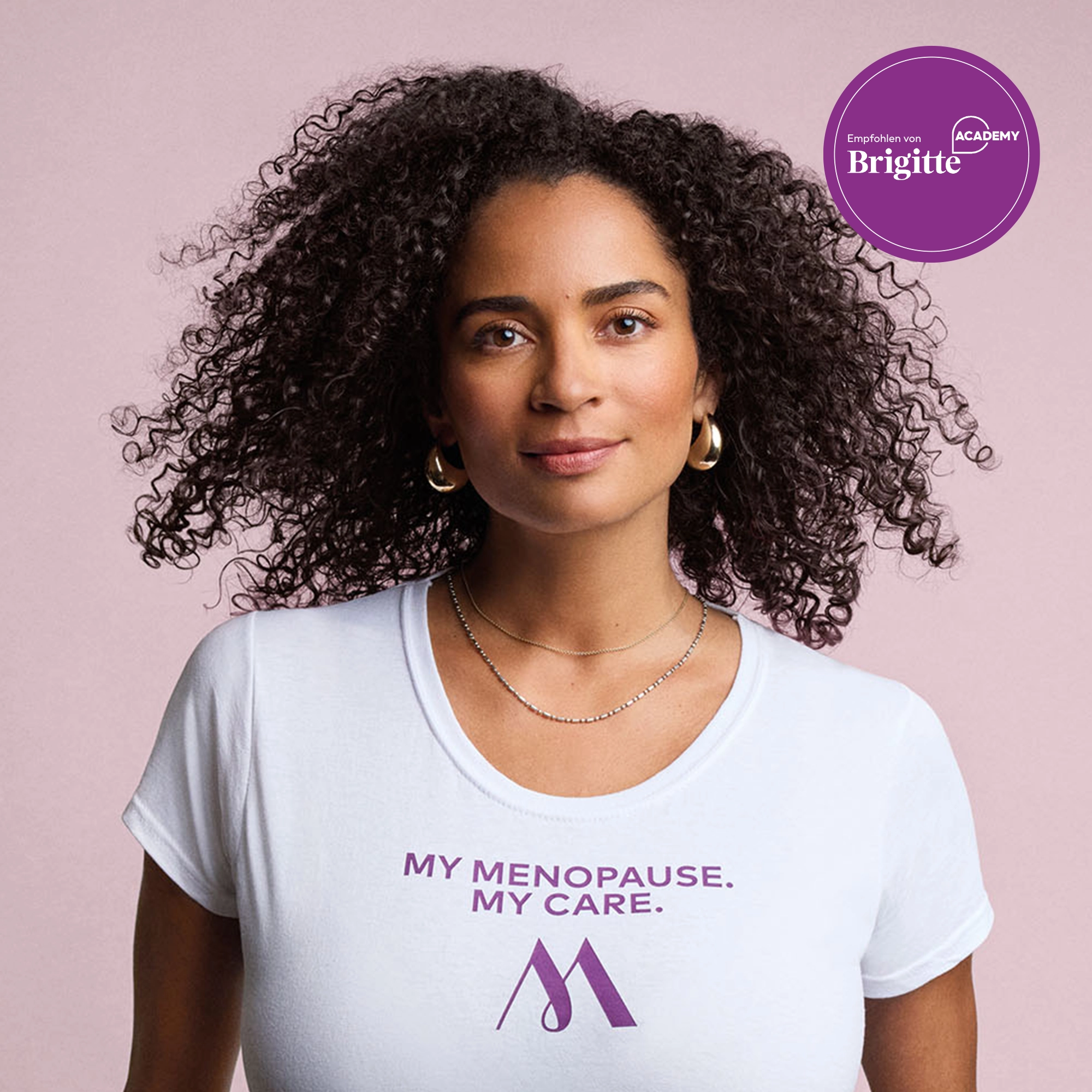 Frau mit lockigem Haar und weißem Shirt mit Aufschrift „MY MENOPAUSE. MY CARE.“ vor rosa Hintergrund