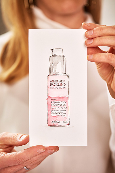 Hand hält eine Zeichnung der ANNEMARIE BÖRLIND Rosenblüten Vitalpflege in Flasche mit Pumpspender, 30 ml.
