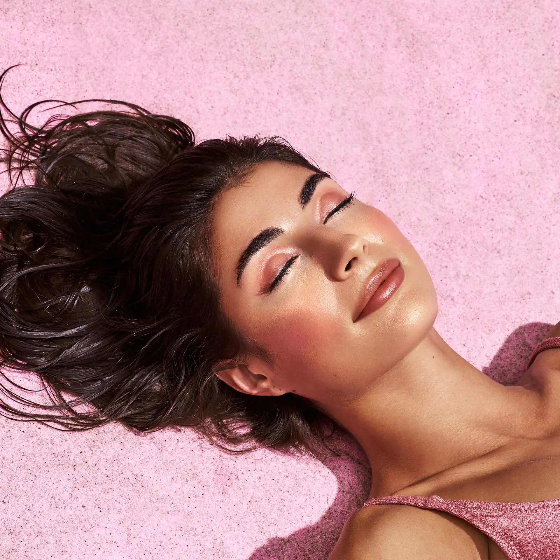 Frau liegt entspannt auf rosa Untergrund, geschlossene Augen, natürlicher Make-up-Look mit warmen Tönen