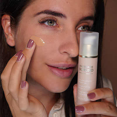 Frau trägt ANNEMARIE BÖRLIND Feuchtigkeitscreme auf Wange auf, hält Produktflasche in der Hand