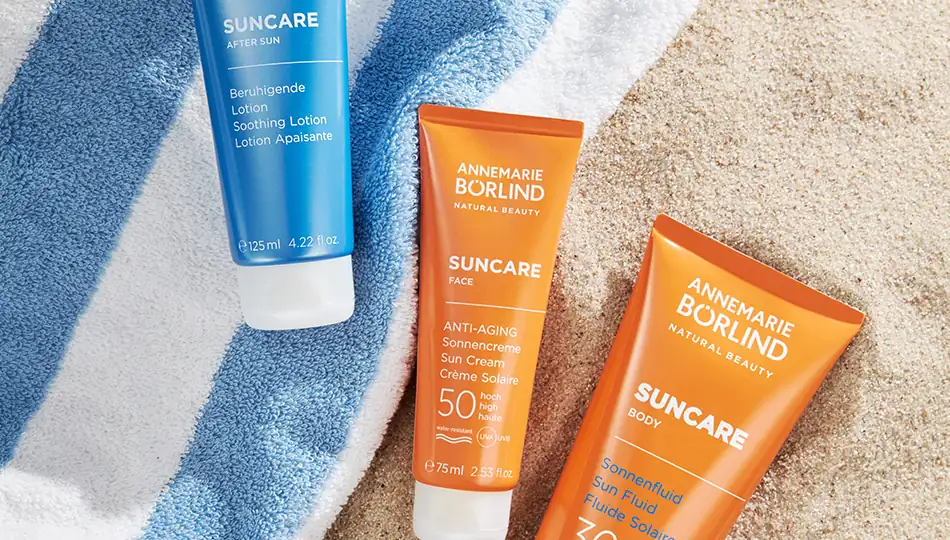 ANNEMARIE BÖRLIND SUNCARE-Produkte auf Handtuch und Sand: After Sun Lotion, Anti-Aging Sonnencreme LSF 50 fürs Gesicht und Sonnenfluid LSF 30 für den Körper.