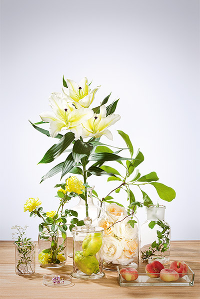 Verschiedene Glasvasen mit weißen Lilien, gelben Rosen, grünen Blättern und Pfirsichen auf Holzfläche