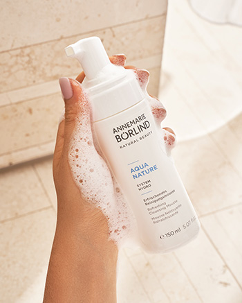 Hand hält ANNE MARIE BÖRLIND AQUA NATURE Reinigungsschaum mit Schaum auf der Haut vor hellem Fliesenhintergrund