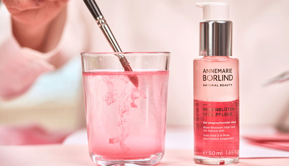 ANNEMARIE BÖRLIND Rosenblüten Vitalpflege für anspruchsvolle Haut neben Glas mit rosa Flüssigkeit und Pinsel
