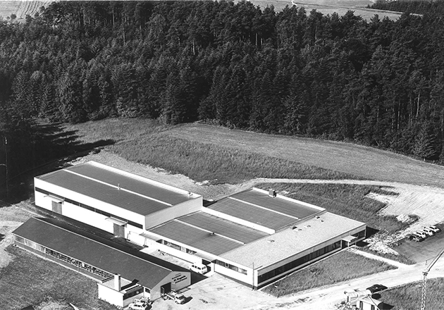 Luftaufnahme eines Industriegebäudes mit mehreren Hallen am Waldrand und angrenzenden Parkplätzen