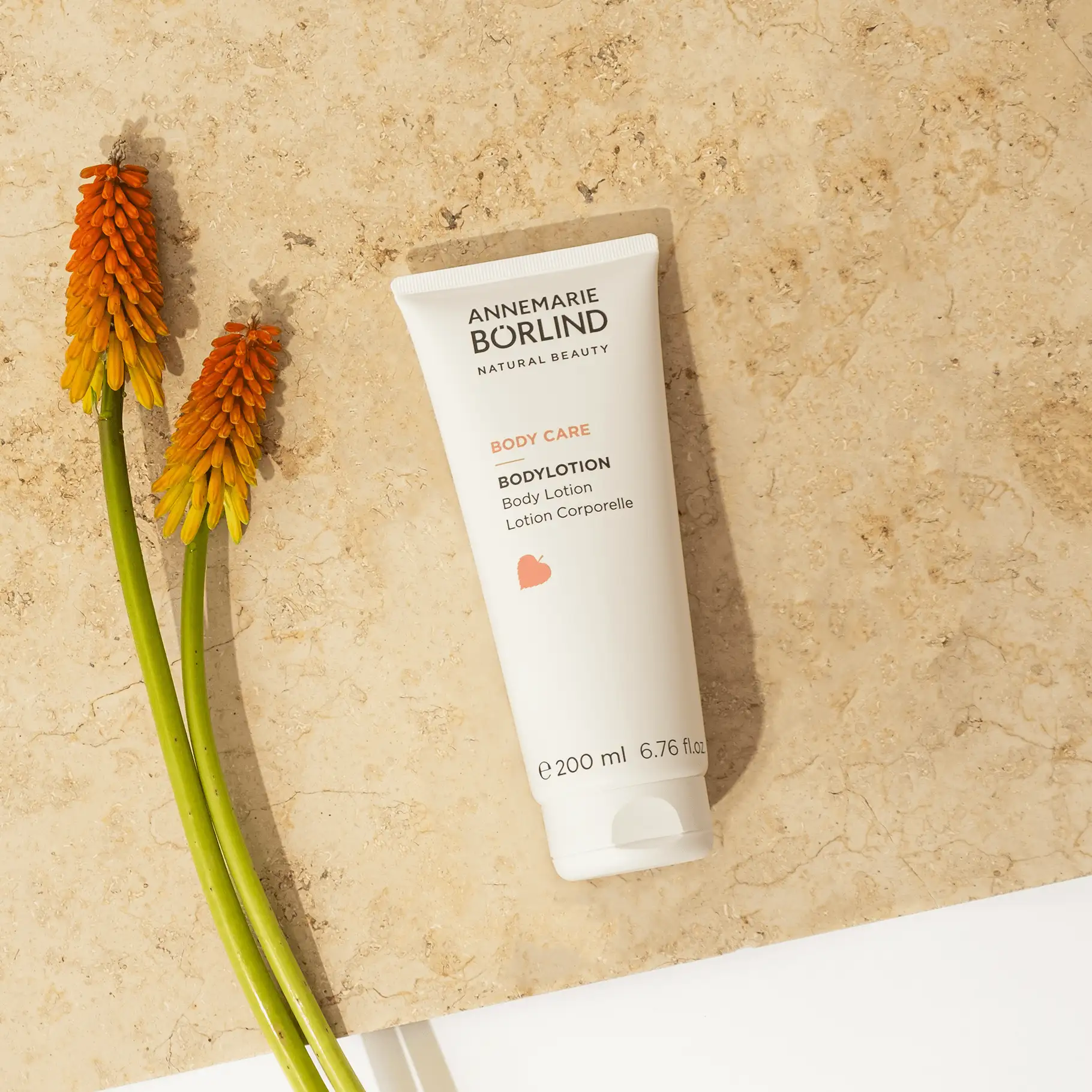 ANNEMARIE BÖRLIND Body Care Bodylotion 200 ml auf hellem Stein mit zwei orangen Blütenstielen