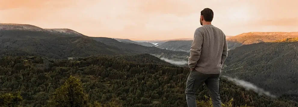 Mann steht auf Berggipfel und blickt auf bewaldete Täler und Hügel im warmen Abendlicht