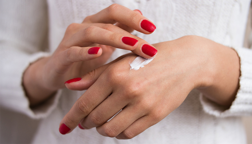 Handcreme wird auf die Handrückseite aufgetragen, Hände mit roten Fingernägeln vor weißem Pullover