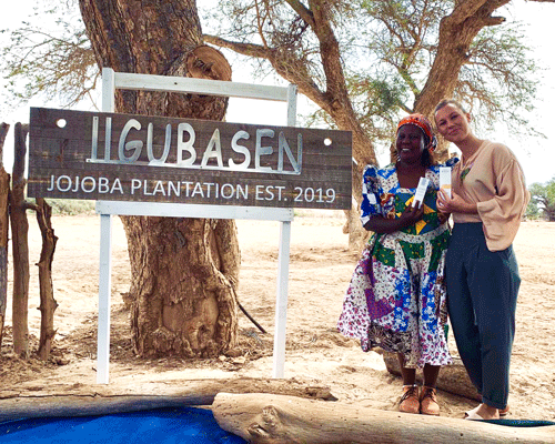 Zwei Frauen stehen neben einem Schild der Jojoba-Plantage Ilgubasen in Namibia, halten Produkte in der Hand.