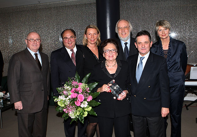 Sechs Personen in formeller Kleidung, Frau mit Blumenstrauß und Auszeichnung in der Mitte, Gruppenfoto bei Veranstaltung