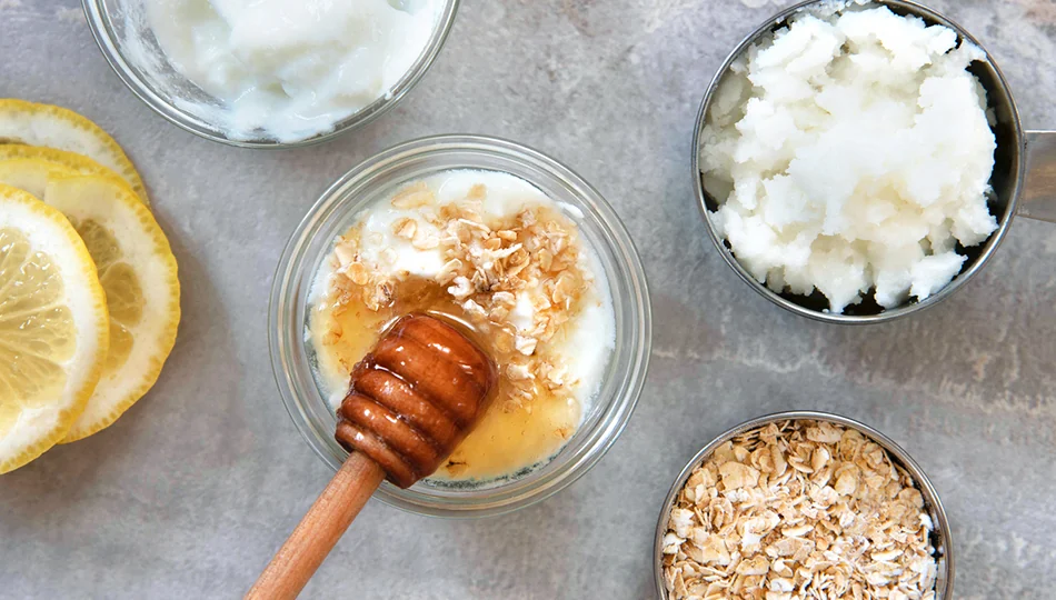 Naturkosmetik Trockene Haut Glas mit Honig, Joghurt und Haferflocken, daneben Kokosöl, Haferflocken und Zitronenscheiben auf grauem Untergrund