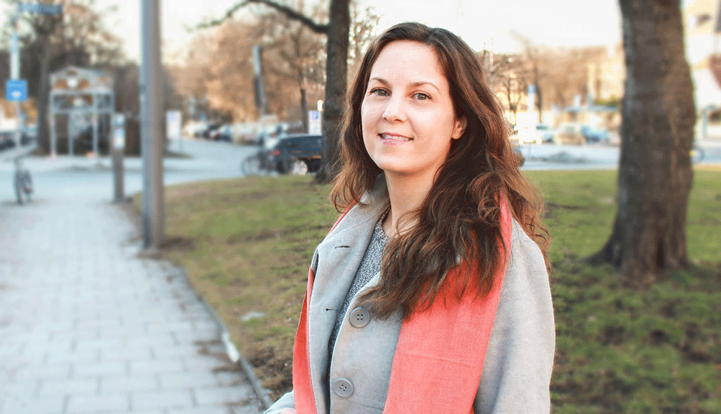 Frau mit langem Haar und grauem Mantel steht lächelnd auf einem Gehweg in einer Stadt bei Tageslicht