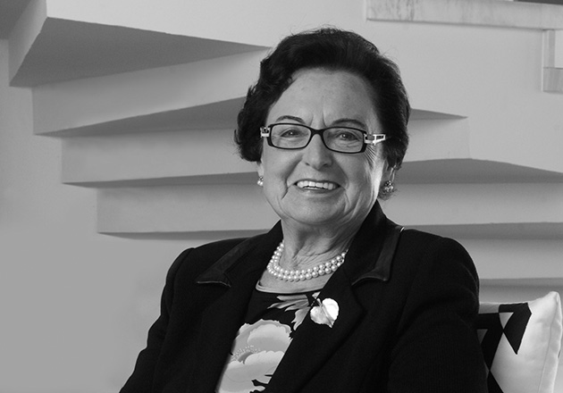 Annemarie Lindner mit Brille und Perlenkette sitzt lächelnd vor einer Treppe in einem Innenraum