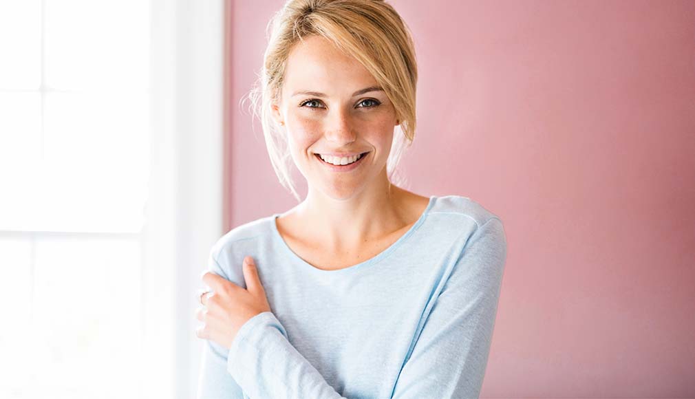 Frau lächelt in hellem Raum, trägt hellblaues Shirt, umarmt sich selbst vor rosa Wand, freundliche Stimmung