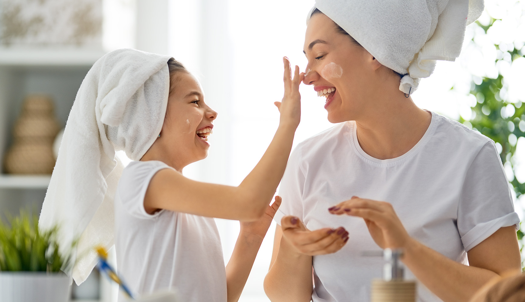 Mädchen trägt Creme auf das Gesicht der Frau auf, beide lachen, Handtücher um die Haare, helle Umgebung
