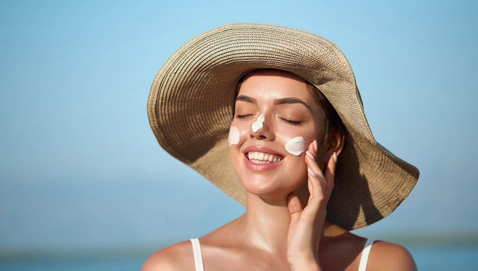 Bräune erhalten Bräune erhalten: Frau mit Sonnenhut trägt lächelnd Sonnencreme auf ihr Gesicht auf, um ihre Sommerbräune zu schützen und die Haut zu pflegen.