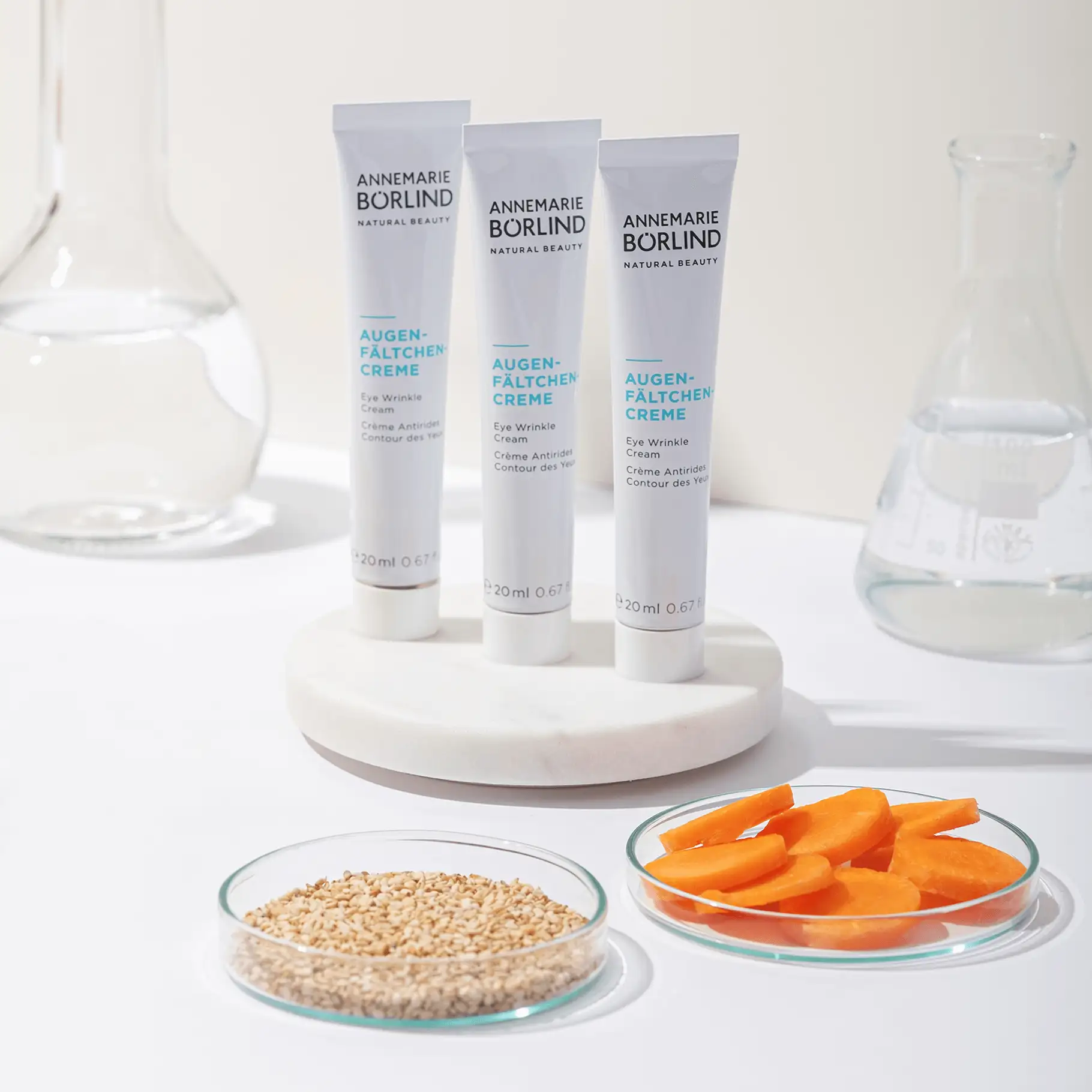 ANNEMARIE BÖRLIND Augen-Fältchen-Creme in drei Tuben mit Sesam und Karottenscheiben auf weißem Hintergrund