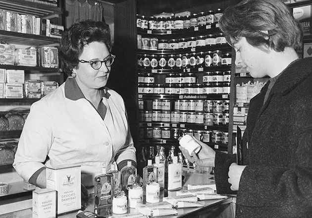 Verkäuferin berät Kundin an Ladentheke mit Kosmetikprodukten in einem Geschäft der 1950er Jahre