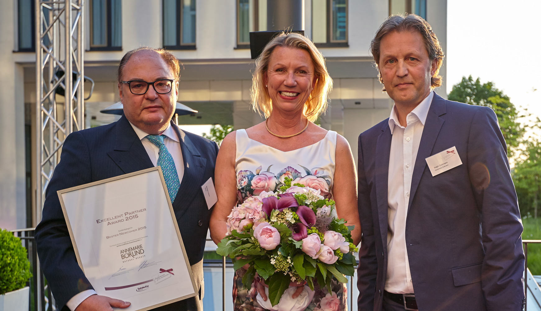 Drei Personen bei Award-Verleihung, Mann hält Urkunde, Frau trägt Blumenstrauß, Abendlicht im Hintergrund